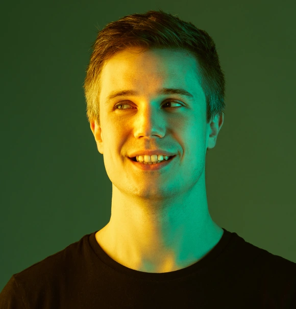 Person with short hair smiling, illuminated by green and yellow lighting, wearing a black shirt, looking upwards.