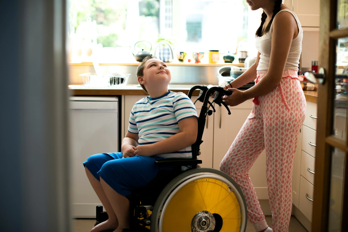 A child in a wheelchair and a woman stand in a sunlit kitchen. The child looks up at the woman, who is holding the wheelchair.