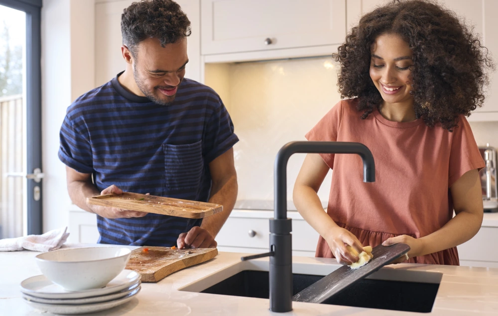 A man and woman clean up in a kitchen; he holds cutting boards, and she scrubs one under a faucet.