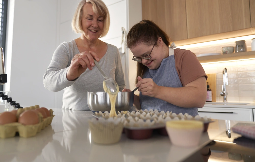 Two women are baking in a kitchen. One is pouring batter into cupcake liners while the other holds a mixing bowl. Eggs and ingredients are visible on the countertop.