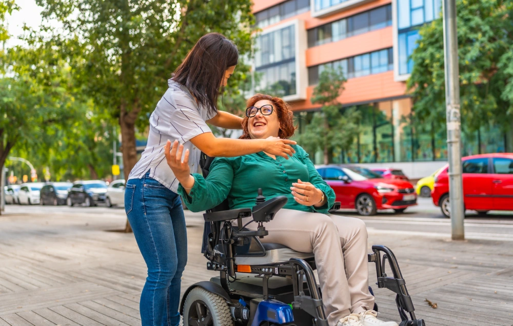 A woman in a wheelchair is assisted by another woman on a city sidewalk. They are smiling, surrounded by trees and parked cars, with a building in the background.