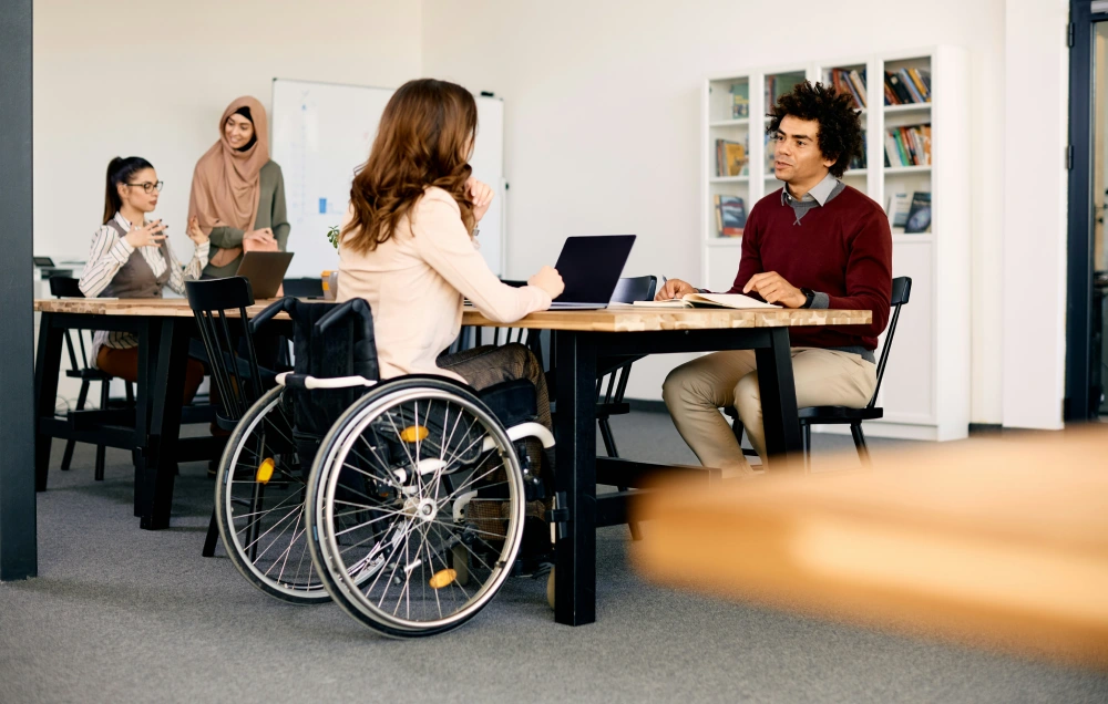 Four people in a meeting room. A woman in a wheelchair and a man with a laptop sit at a wooden table, while two others, a woman standing and another seated, engage in conversation in the background.