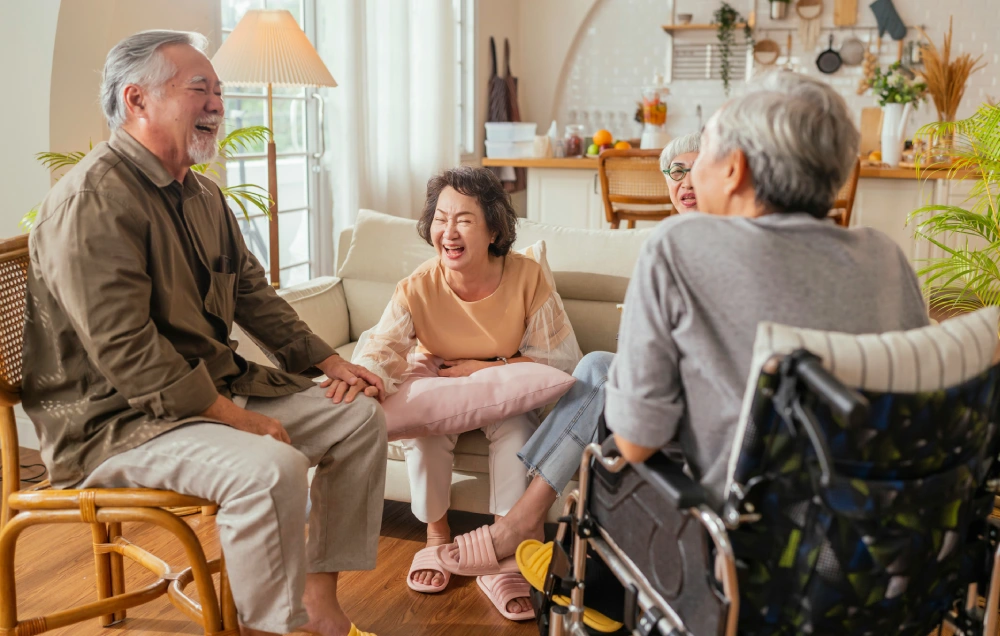 Four older adults are sitting and smiling in a cozy living room. One person is in a wheelchair. The room has warm lighting and a tidy kitchen is visible in the background.