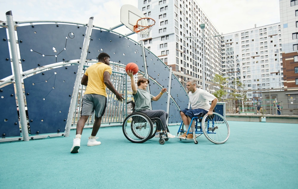 Three men, two in wheelchairs, play basketball on an outdoor court. One man has the ball, aiming to pass. Tall buildings are visible in the background.