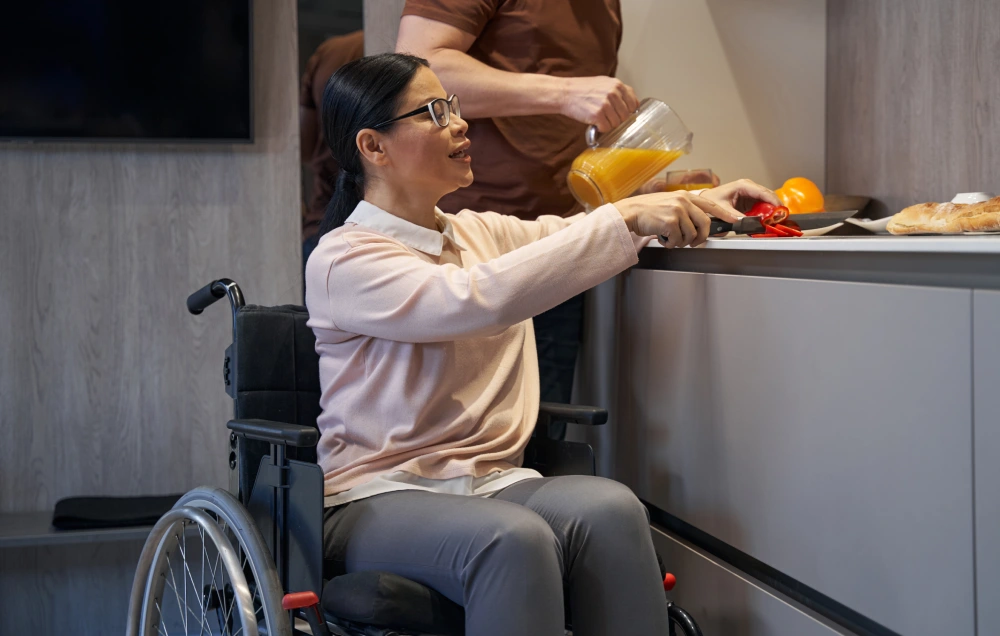 A woman in a wheelchair reaches for a plate on a counter, while a person next to her pours orange juice from a jug.