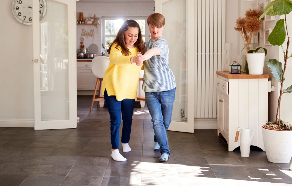 Two people indoors, joyfully dancing together in a spacious, well-lit room with wooden furniture and potted plants.