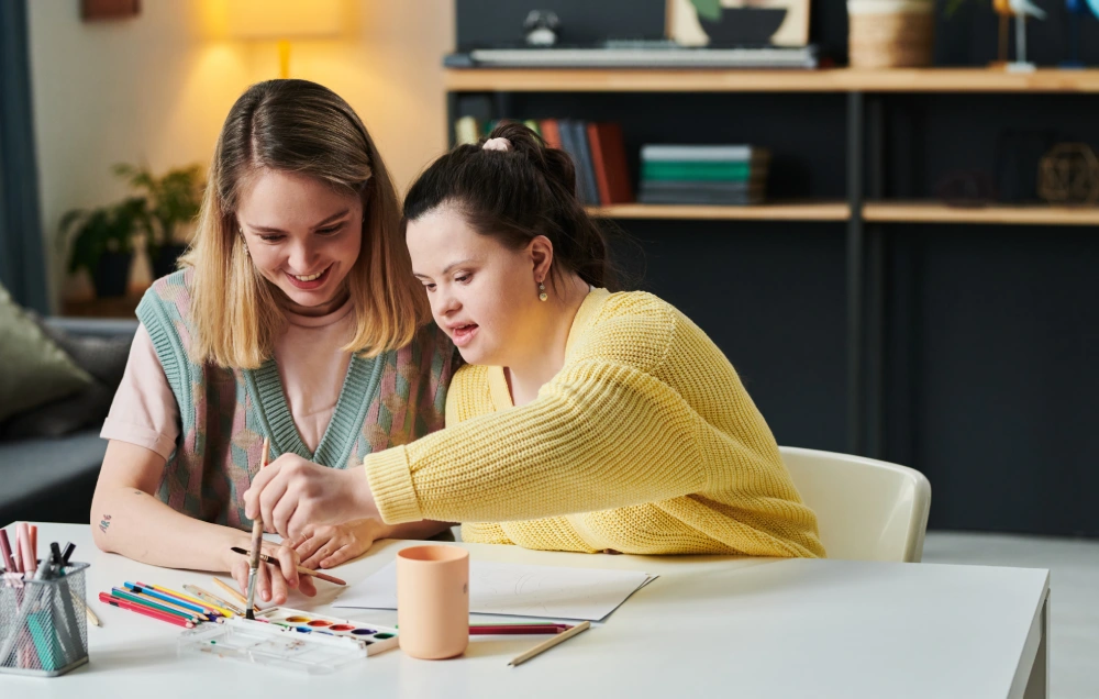 Two people sitting at a table engaging in an art activity with paints and colored pencils. One person assists the other, who is holding a paintbrush. Bookshelves are in the background.