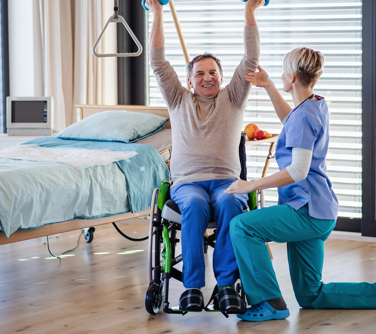 A person in a wheelchair lifting dumbbells overhead with the assistance of a healthcare worker in a brightly lit room.